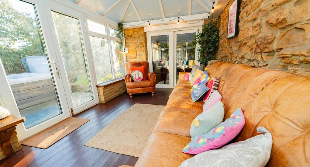 A sun-drenched social hub in the Tranquil Escape conservatory, showcasing the large leather couch and patio doors that lead directly to the hot tub and private garden oasis.