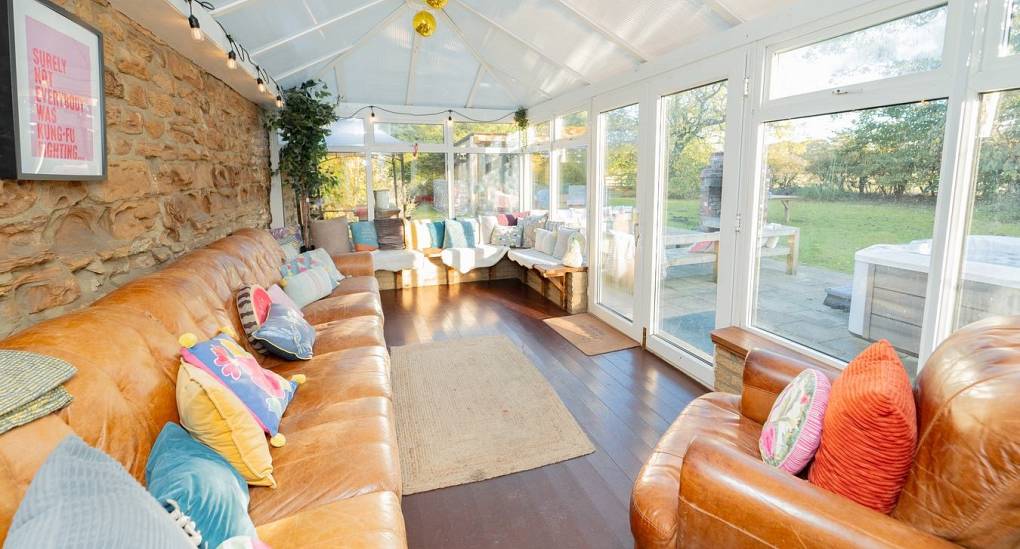 The bright and airy conservatory at Tranquil Escape, a rural Newcastle hen do house, featuring a massive leather social sofa, festoon lighting, and a traditional exposed stone wall.