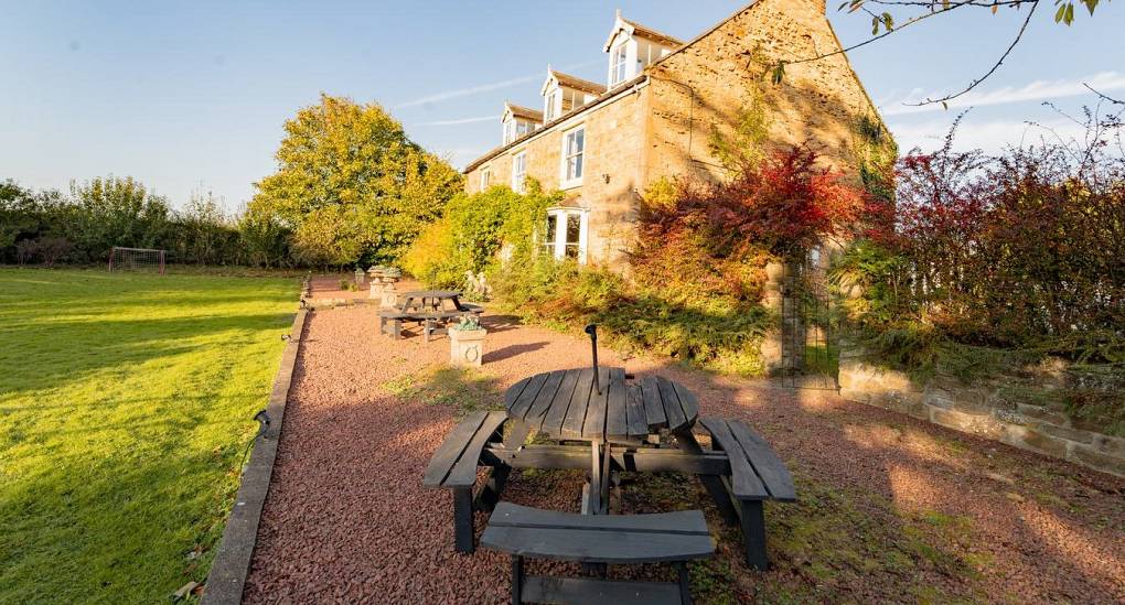 Side view of the traditional stone manor at Tranquil Escape, showing the red gravel patio area with picnic benches and the secluded, leafy surroundings of this detached estate.